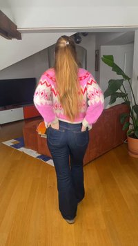 Άτομο που στέκεται μέσα σε εσωτερικό χώρο με μακριά ξανθά μαλλιά, φοράει ροζ πουλόβερ με σχέδια και σκούρο τζιν, τα χέρια πίσω στις τσέπες, κοντά σε φυτό και καναπέ.