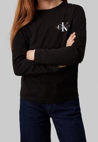 Černé tričko s dlouhým rukávem vyrobené z bavlny. Na hrudi má bílé logo Calvin Klein. Kombinováno s tmavými džíny.