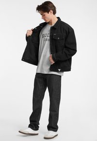 Juodas džinsinis švarkas su mygtukų detalėmis, pilkas marškinėliai su "GUESS JEANS" tekstu, tamsūs džinsai ir balti sportbačiai su pilkais akcentais.