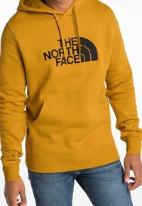 Žlutá mikina s kapucí vyrobená z bavlněné směsi, má černé logo, klokánkovou kapsu, žebrované manžety a lem, navržena pro volnočasové nošení.