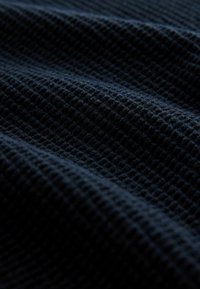 Strukturierter marineblauer Stoff mit einem Rautenmuster, das ausgeprägte Erhebungen und Vertiefungen zeigt und ein reichhaltiges, haptisches Erscheinungsbild erzeugt.