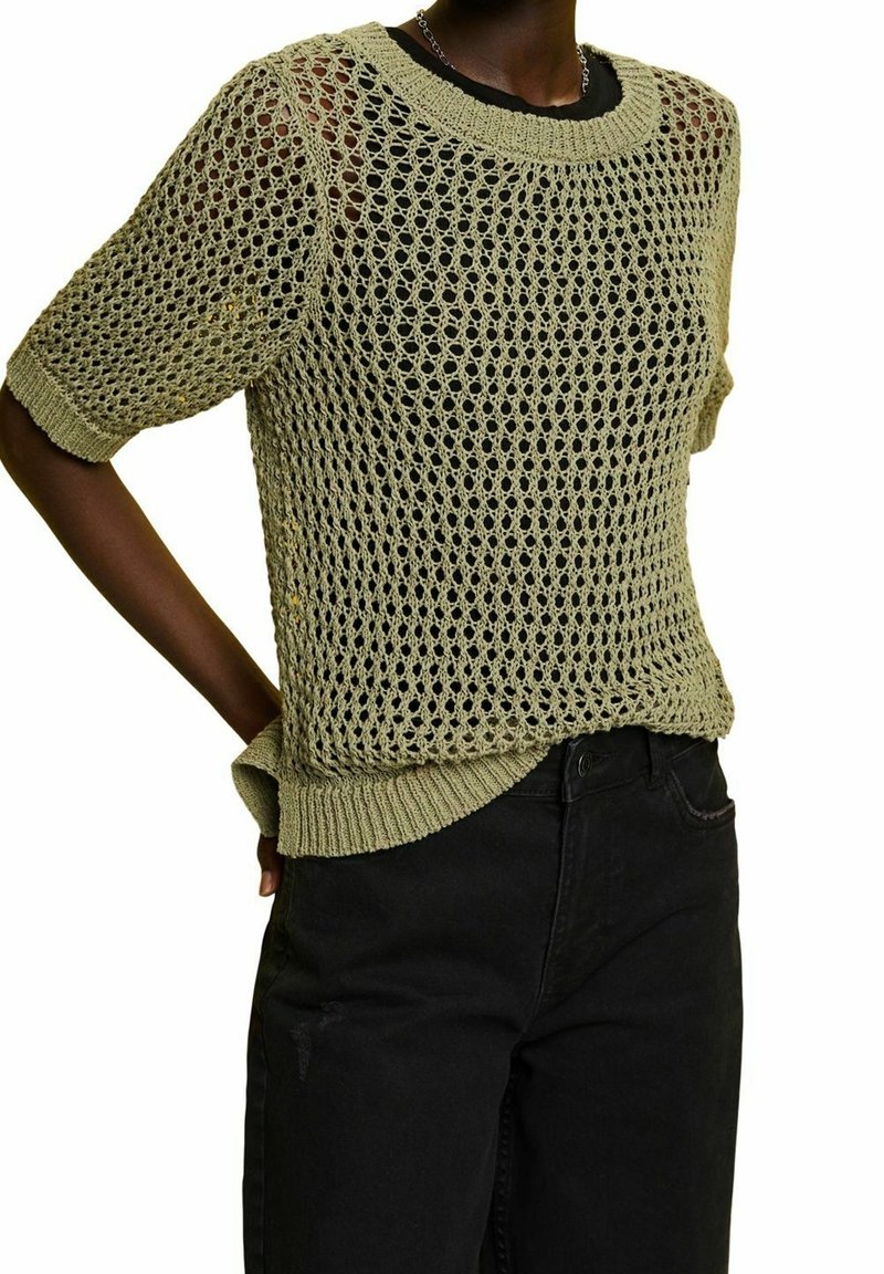 Personne portant un pull beige en maille ajourée à manches courtes par-dessus un haut noir et un pantalon noir taille haute, avec une main derrière le dos.