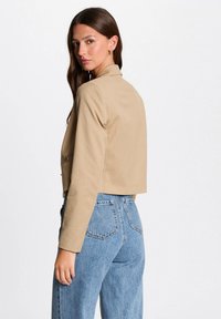 Korte beige gestreepte jas met lange mouwen, voorzien van een geknipt kraagje en knoopaccenten, gecombineerd met hoge taille blauwe spijkerbroek.