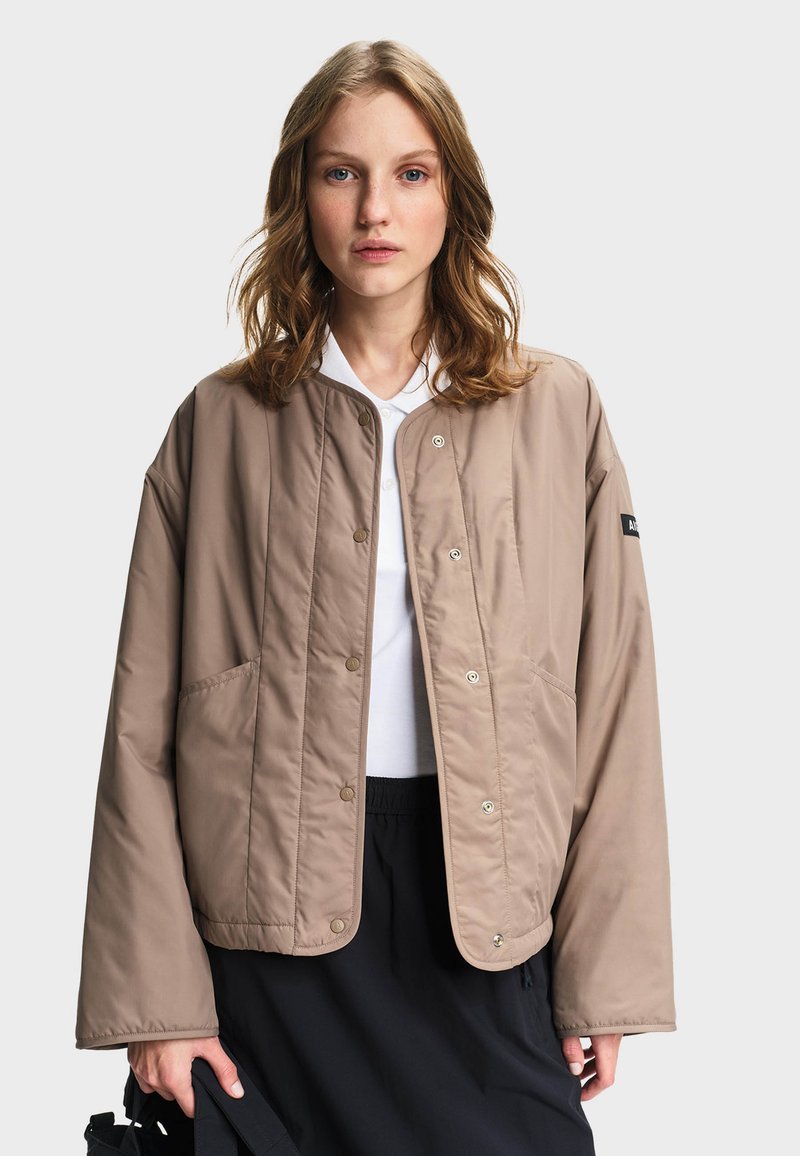 Femme aux cheveux blonds ondulés portant une veste beige boutonnée sur une chemise blanche et une jupe noire, tenant un sac noir sur un fond uni.