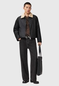 Chaqueta bomber negra con cuello de pelo sintético, superpuesta sobre una camiseta de punto oscura con estampado, combinada con unos pantalones vaqueros anchos negros y un bolso tote negro.