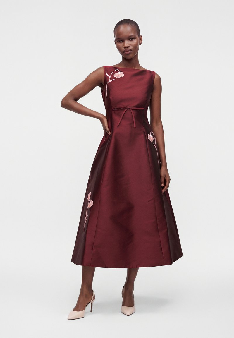 Femme portant une robe midi bordeaux sans manches avec broderie florale rose, posant une main sur la hanche, en escarpins clairs.