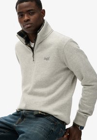 Szary sweatshirt na zamek z wysokim kołnierzem i subtelnym logo, noszony na niebieskiej i żółtej koszuli w kratę, w połączeniu z dżinsami.