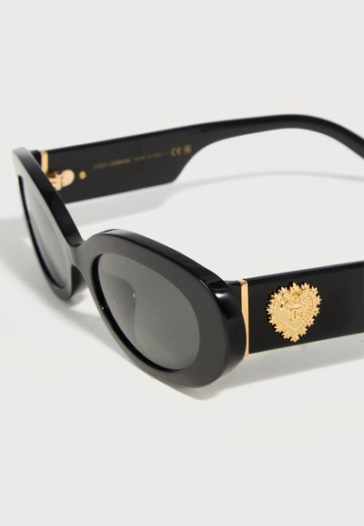 Μαύρα οβάλ γυαλιά ηλίου με σκουρόχρωμους φακούς και χρυσό έμβλημα σε σχήμα καρδιάς στο πλάι, με περίτεχνη λεπτομέρεια και αρχικά "DG".