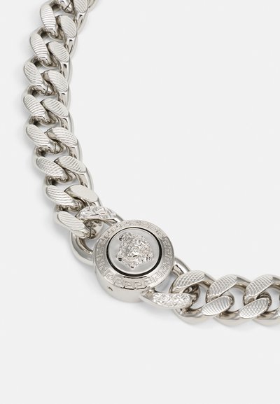 Srebrna verižica z debelimi povezavami z teksturiranimi poudarki, okroglo zapiranje z levjim emblemom in zapletenimi detajli okoli zapiranja.