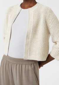 Cardigan en tricot crème à texture, avec un col rond et un devant ouvert, associé à un débardeur côtelé gris clair et un pantalon beige à taille élastique.