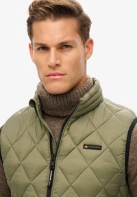 Groene gewatteerde vest met een hoge kraag, ritsluiting en zwarte accenten; heeft een logo-opdruk op de borst en een zachte bruine turtleneck eronder.