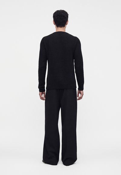Mann steht mit dem Rücken zur Kamera, trägt ein eng anliegendes schwarzes Langarmshirt und lockere schwarze weite Hosen vor einem schlichten Hintergrund.