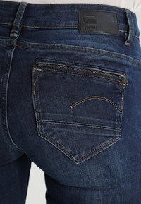 Los vaqueros de mezclilla azul oscuro cuentan con un bolsillo trasero con cierre de cremallera, costuras reforzadas y un parche de cuero de la marca cerca de la cintura.