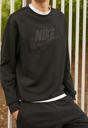 Person trägt schwarzen Nike-Sweatshirt und passende Hose, steht im Freien nahe weißen Metallstangen mit Grünpflanzen im Hintergrund.