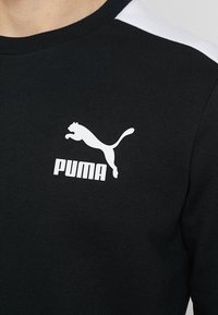 Černá mikina s bílými akcenty na ramenou, s výrazným bílým logem Puma a hladkou, vroubkovanou texturou látky.
