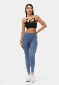 Melnas sporta krūšturis ar krustveida siksnām, apvienots ar augsto jostasvietu ziliem legingiem. Modelis valkā baltas sporta kurpes, stāvot pret vienkāršu fona.