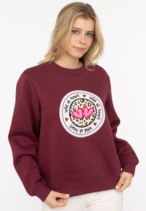 Bordeauxrotem Sweatshirt mit einem runden Grafikdesign mit Leopardenmuster und pinken Herzen, verziert mit dem Satz "wild at heart" in Schwarz.