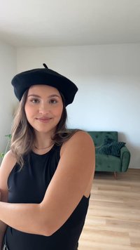 Černý baret s malým horním poutkem, který nosí osoba v bezrukávovém černém topu. V pozadí je zelená pohovka a světlá dřevěná podlaha.