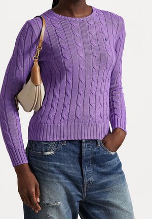 Personne portant un pull torsadé violet, un jean bleu délavé et un petit sac à main beige avec des détails en cuir marron.