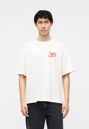 Jonge man draagt een wit oversized T-shirt met een klein rood "Castrol"-logo en een autografiek op de borst, gecombineerd met een zwarte broek.