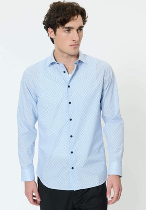 Ung mand med krøllet hår iført en lyseblå, mønstret skjorte med knapper og sorte bukser, der kigger mod højre mod en hvid baggrund.