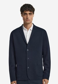 Homme portant un blazer boutonné bleu marine foncé sur une chemise blanche, se tenant devant un fond blanc uni, tourné vers l'avant.