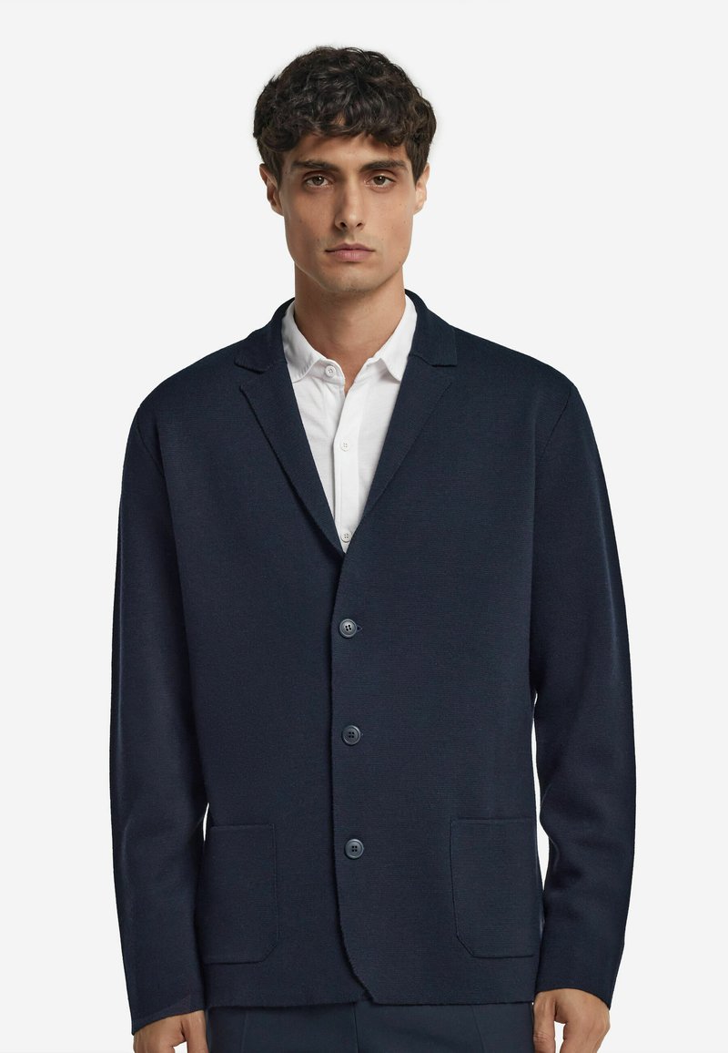 Homme portant un blazer boutonné bleu marine foncé sur une chemise blanche, se tenant devant un fond blanc uni, tourné vers l'avant.