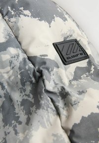 Tissu à motifs gris et blanc avec une texture et un patch en caoutchouc noir portant un logo avec des designs géométriques et des coordonnées.