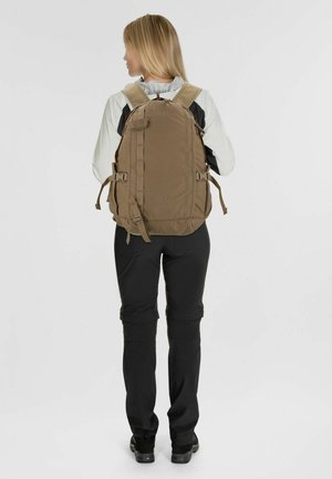 Zaino khaki con spallacci imbottiti, multiple cinghie, due tasche laterali e una finitura strutturata, indossato da una persona con pantaloni neri.