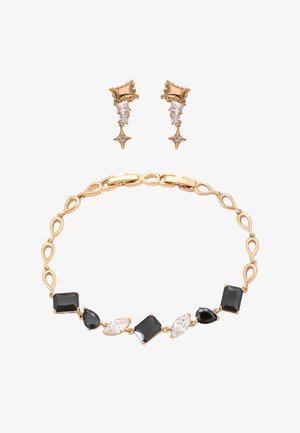Złota bransoletka z naprzemiennie ułożonymi czarnymi i przezroczystymi geometrycznymi kamieniami ozdobnymi, zestawiona z złotymi kolczykami z gwiazdkowymi i przezroczystymi akcentami kamieni.