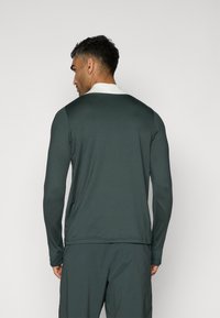 Cămașă cu mâneci lungi din țesătură verde închis, cu guler alb. Prezintă o croială lejeră și o textură netedă, concepută pentru confort.