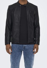 Zwarte leren jas met een opstaande kraag, ritssluiting en zijzakken. Draagt over een zwart shirt en blauwe spijkerbroek. Soepele textuur.