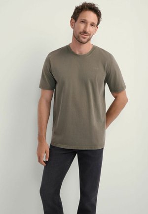 Mężczyzna z krótkimi kręconymi włosami nosi oliwkową koszulkę z krótkim rękawem i czarne dżinsy, stojąc na tle jednolitego, jasnobeżowego tła.