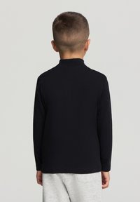 Koszula z długim rękawem z jednolitej czarnej tkaniny, z wysokim kołnierzem, gładką fakturą i prostym krojem, noszona z szarymi spodniami.