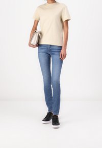 Beige bomull t-shirt, slim blå jeans och svarta sneakers med vita sulor. En ljusgrå clutchväska hålls i vänster hand.