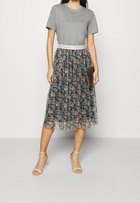 Kobieta ubrana w szare t-shirt, kwiecistą plisowaną midi spódnicę z paskiem "Dreams Happen", w cielistych sandałach na kostkę, trzymająca ciemną kopertówkę.