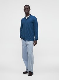 Homme debout portant une chemise bleue texturée à boutons, un t-shirt blanc, un jean large bleu clair et des chaussures marron foncé sur un fond uni.