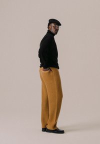 Schwarzer Rollkragenpullover, locker sitzende, strukturiert senfgelbe Hose, schwarze Schirmmütze und schwarze Schuhe. Schlichtes, modernes Design mit minimalen Akzenten.