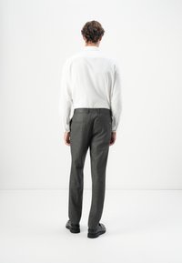 Homme de dos portant une chemise blanche à manches longues, un pantalon habillé gris et des chaussures habillées noires sur un fond blanc.