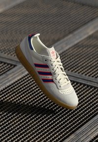 Sapatilha branca com riscas vermelhas e azuis, parte superior em couro texturizado, sola de borracha de goma e detalhes de bico perfurados. Design atlético clássico.