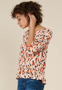 Blouse pêche avec un motif floral noir, manches trois-quarts et un ourlet à volants. Portée avec un jean bleu. Texture de tissu lisse.