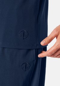 Marineblauwe sportkleding met ribbeltextuur, geborduurd logo en zijvak. Een hand wijst naar het naaddetail van het kledingstuk.