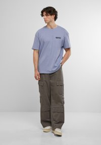 Tricou oversized de culoare violet deschis, cu grafică pe față, asortat cu pantaloni cargo lejeri de culoare gri închis, prevăzuți cu multiple buzunare și o textură a țesăturii netedă.