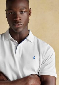 Balts polo krekls ar gaiši ziliem akcentiem uz apkakles un mazu zilu logotipu uz krūts. Materiāls izskatās teksturēts un ikdienišķs.