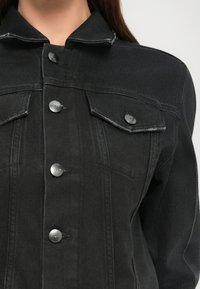 Prim-plan al unei jachete din denim negru cu fața cu nasturi, două buzunare pe piept cu clapete și cusături vizibile pe umeri și mâneci.