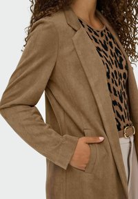 Blazer de pana marrón con un diseño ajustado, solapas de muesca y bolsillos laterales, combinado con una blusa de estampado de leopardo y pantalones beige.