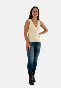 Top sin mangas de color crema con detalles de botones y escote en V, combinado con jeans ajustados de denim y botas de tacón negro hasta la rodilla.