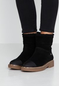 Svarta mocka ankelskor med bruna gummisulor, runda förstärkta tår och fluffig foder, som bärs med svarta tajta byxor.