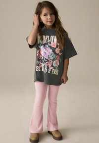 T-shirt ample gris avec un design floral et le texte "SOIS SAUVAGE ET LIBRE", associé à un pantalon flare côtelé rose clair et des chaussures marron.