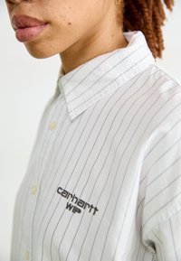 Bijela košulja s dugim rukavima i uskim sivim prugama, s mekanim ovratnikom i teksturiranom tkaninom. Izvezeni logotip "Carhartt WIP" na prsima.